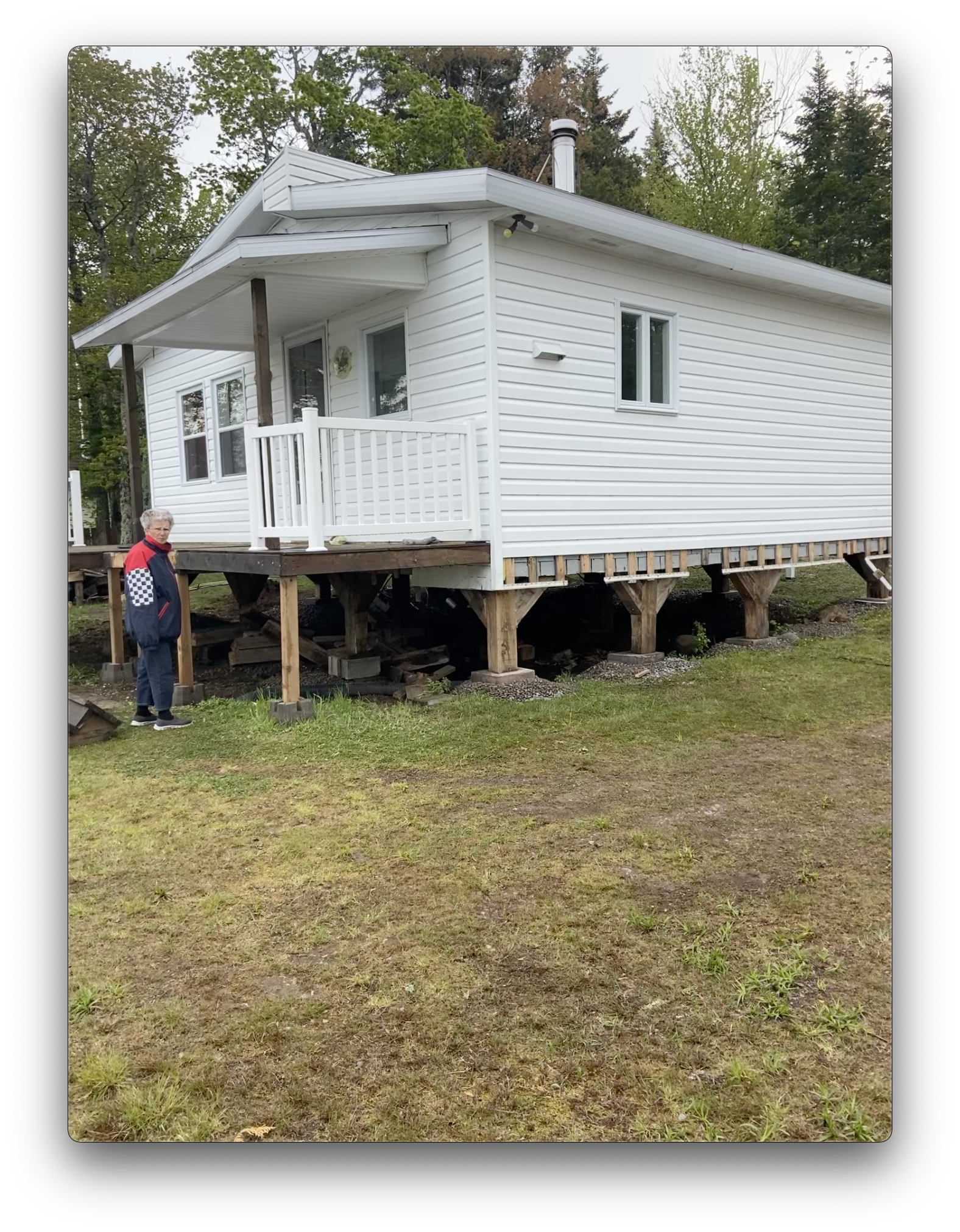 Une personne se tient à côté d'une cabane blanche surélevée sur pilotis en bois, dans une zone herbeuse et boisée.