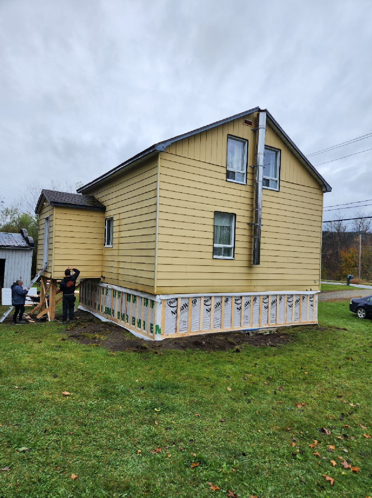 Des ouvriers installent un revêtement extérieur sur une maison jaune à deux étages dont l'isolation des fondations est apparente, par temps nuageux.