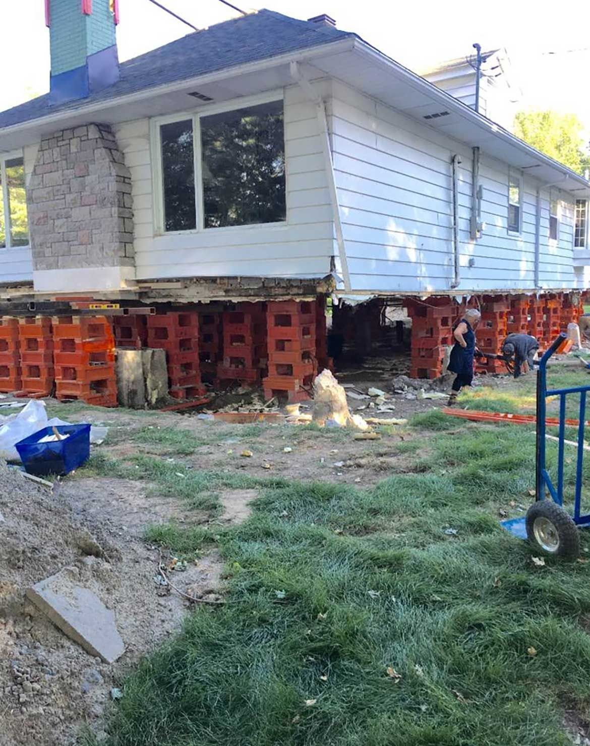 Maison surélevée sur des blocs orange ; ouvriers au sol. Vue extérieure avec herbe et outils.