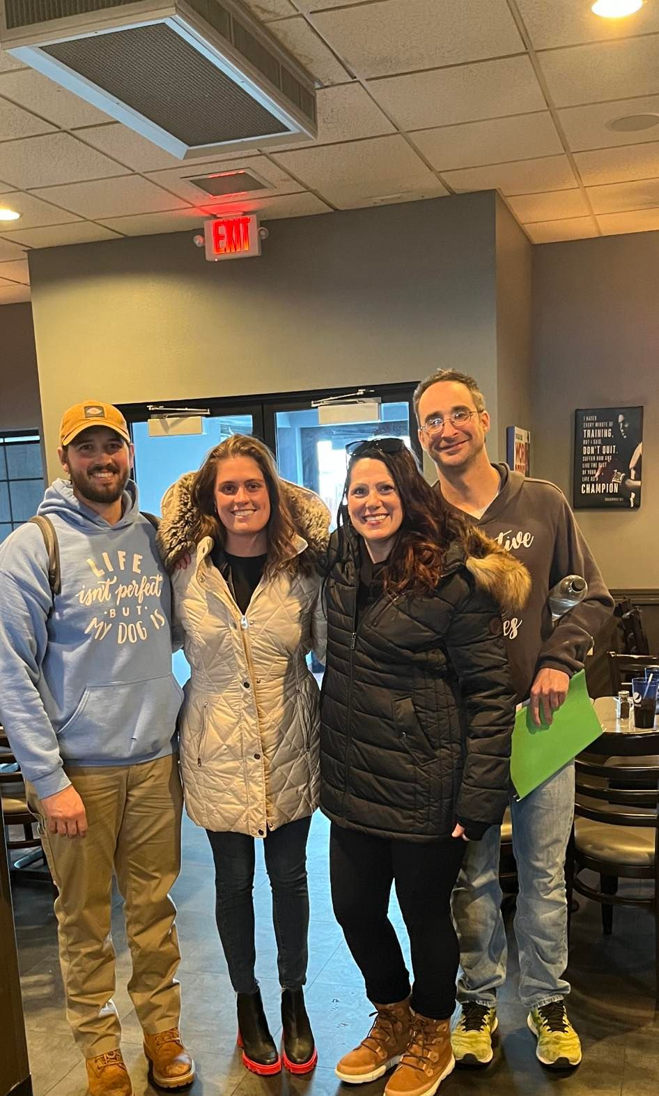 Four people standing, arm in arm, indoors. Two women in coats, two men in a hoodie and a shirt.