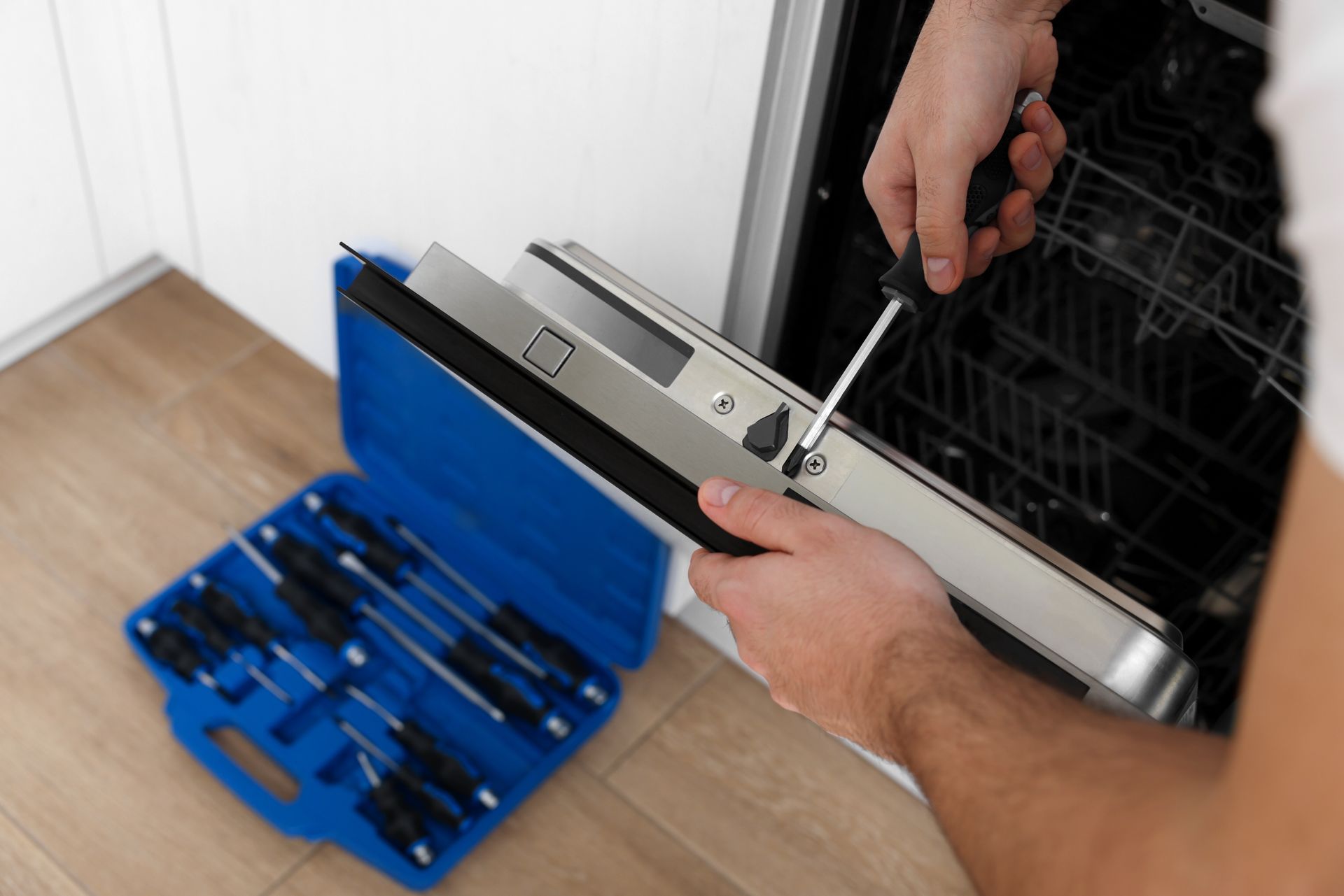 Hands using a screwdriver to adjust a dishwasher door hinge, with a blue tool case on the floor.