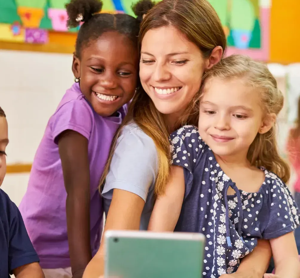 A teacher is sitting next to a group of children looking at a tablet.