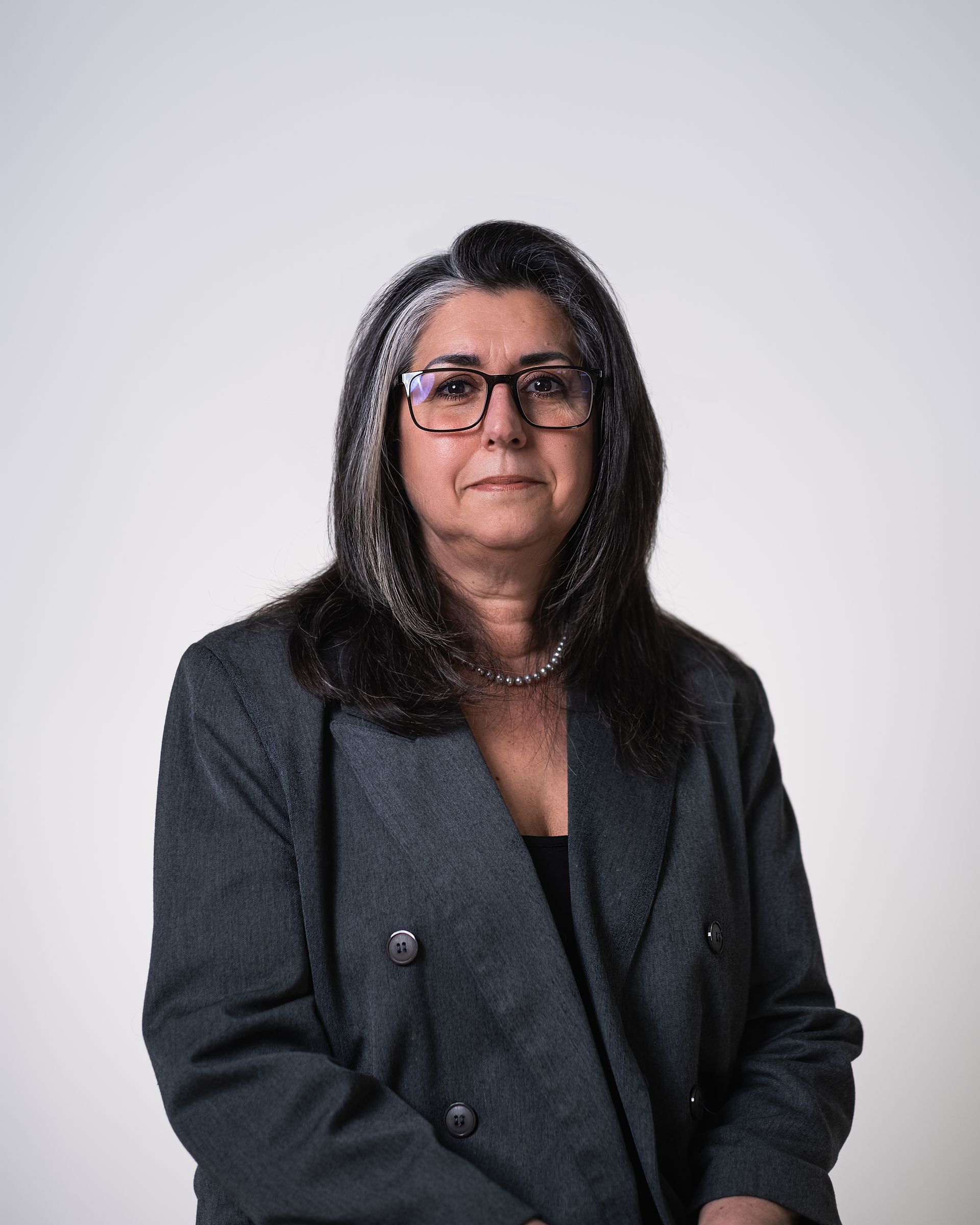 Une femme portant des lunettes et une veste grise se tient devant un fond blanc.