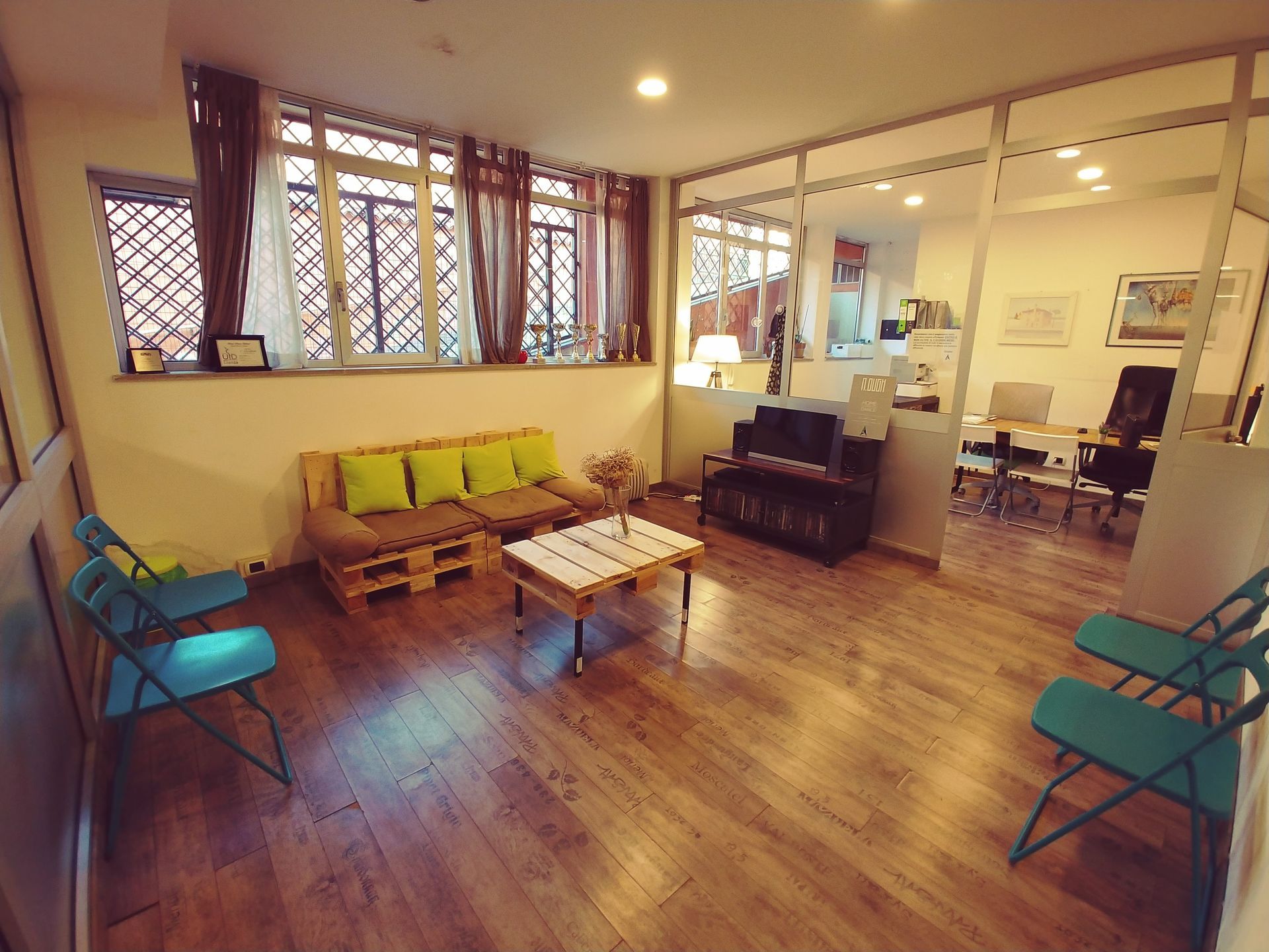 Una sala d'attesa con pavimenti in legno, un divano in pallet, sedie color verde acqua e un'area ufficio con pareti in vetro.
