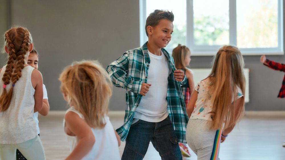 Bambini in una scuola di danza, un ragazzo con una camicia a quadri verde sorride mentre balla con gli altri.