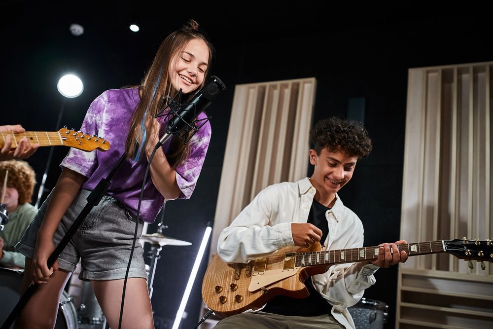 Gruppo di adolescenti in uno studio. Cantante con microfono, chitarrista che suona, batterista sullo sfondo.