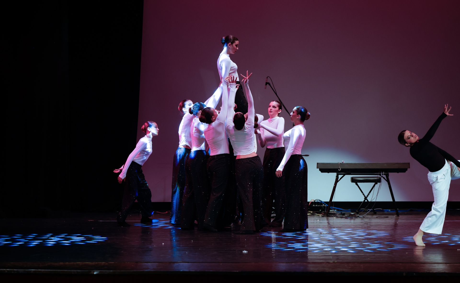 I ballerini si esibiscono sul palco. Uno è sollevato sopra gli altri, con le braccia tese. Altri indossano camicie bianche e gonne scure. La tastiera è sullo sfondo.