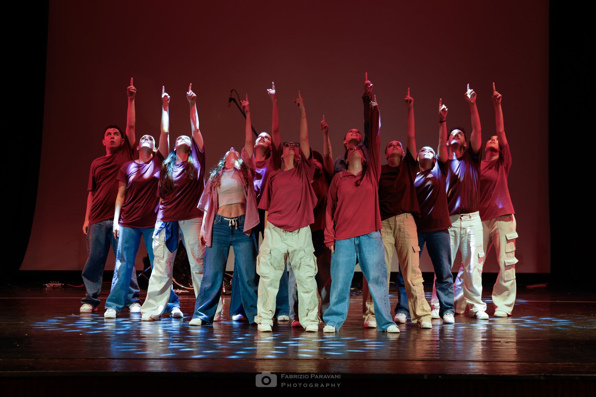 Un gruppo di ballerini si esibisce sul palco indossando abiti coordinati, formando una piramide. Sfondo viola.
