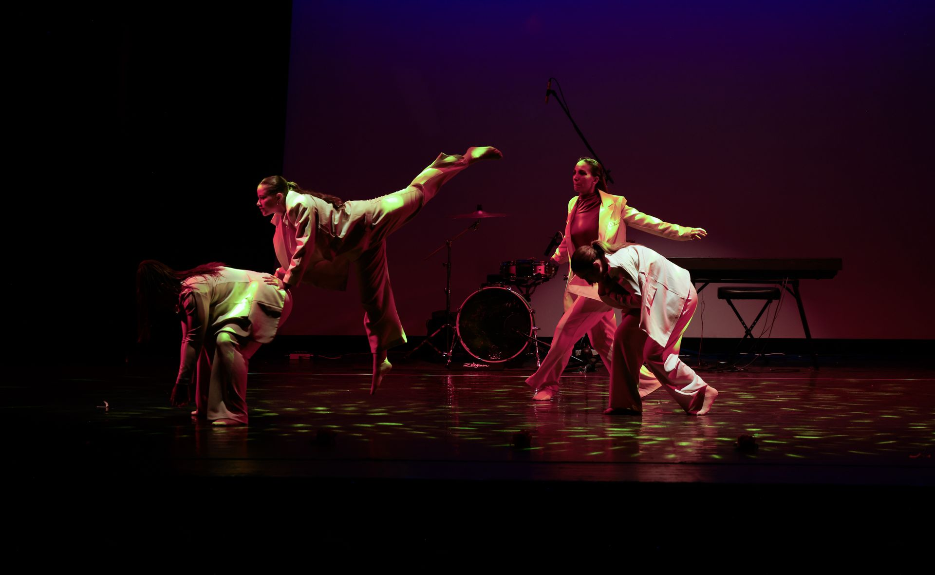 I ballerini sul palco si esibiscono sotto luci colorate, con una batteria e una tastiera sullo sfondo.