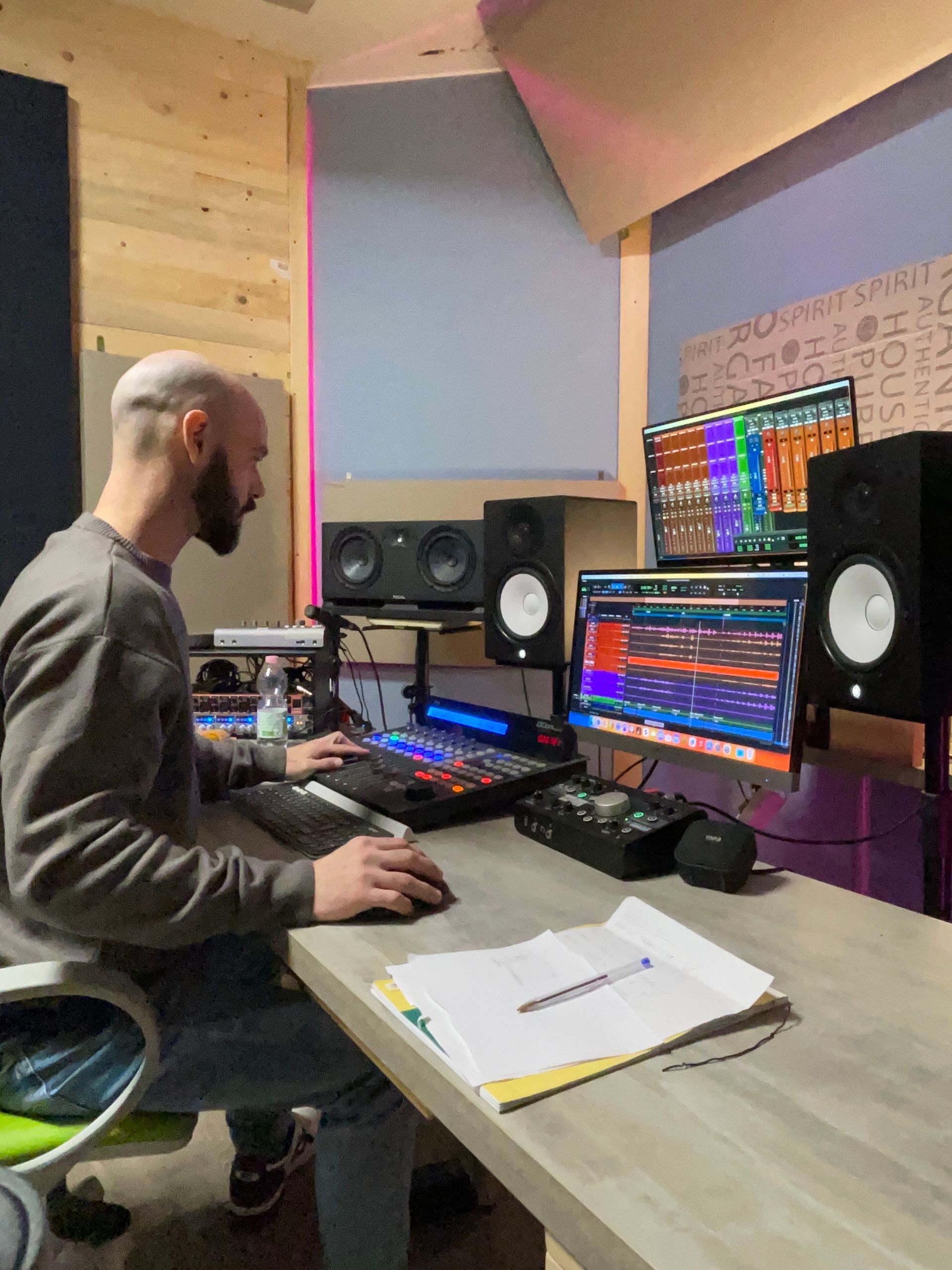 Uomo in uno studio di registrazione che lavora alla produzione audio; i monitor mostrano le forme d'onda, la console di missaggio e i monitor da studio.