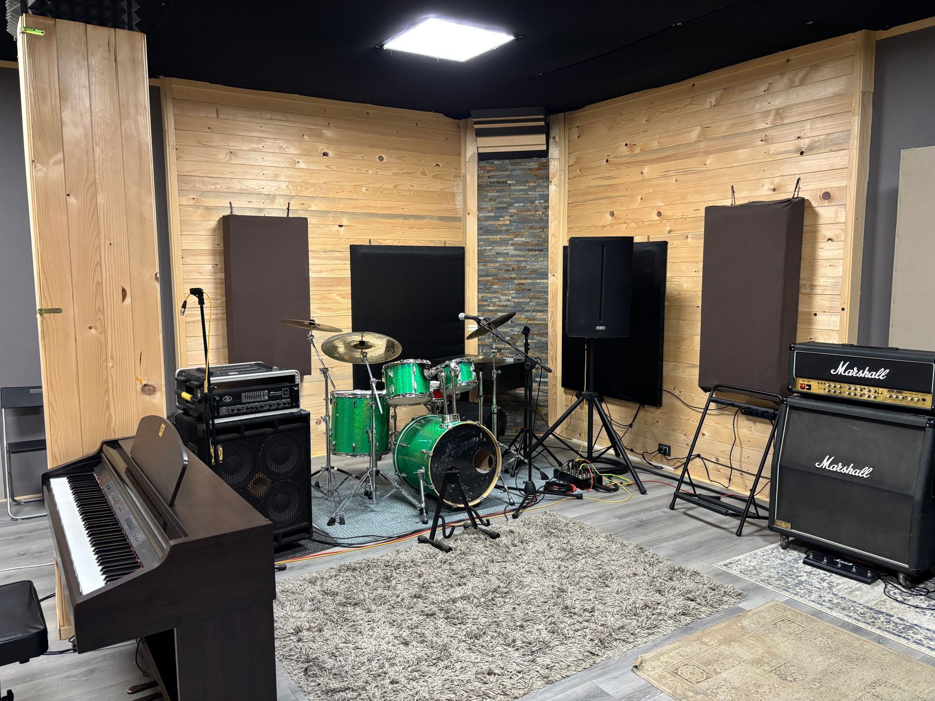Studio musicale con batteria, pianoforte e amplificatori. Attrezzatura marrone, nera e verde. Pareti rivestite in legno.