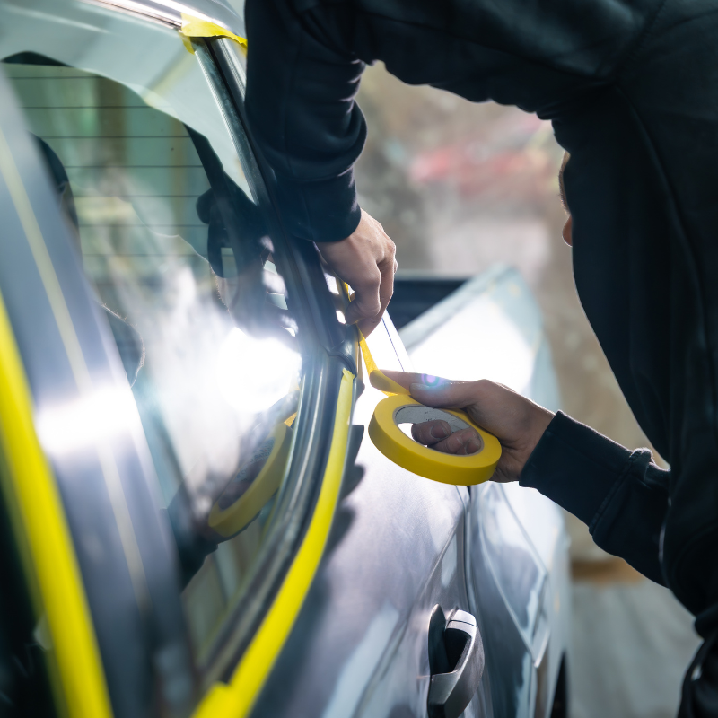 Two people applying yellow tape to a car window in a workshop
