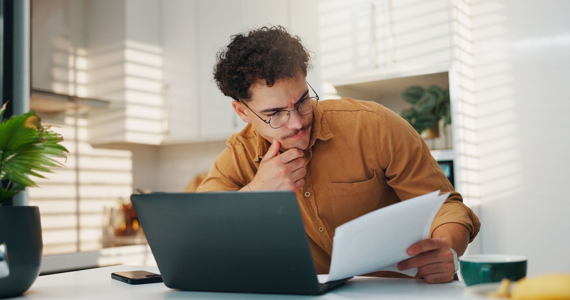 Man with glasses, looking at documents, thoughtful expression, laptop on desk. Kitchen setting. Man with glasses, looking at documents, thoughtful expression, laptop on desk. Kitchen setting.