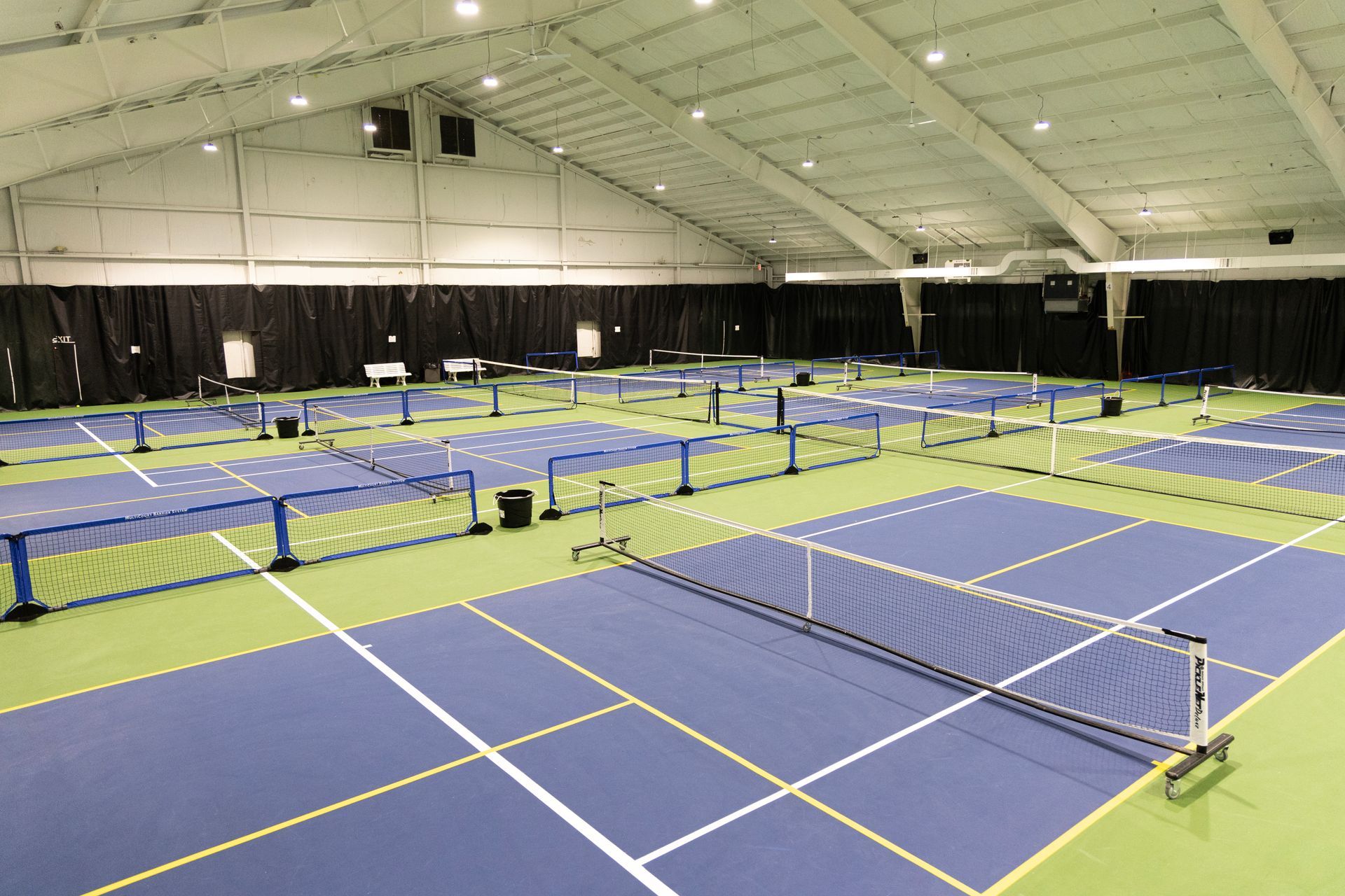 Indoor pickleball court with multiple blue courts, green borders, and black curtains.