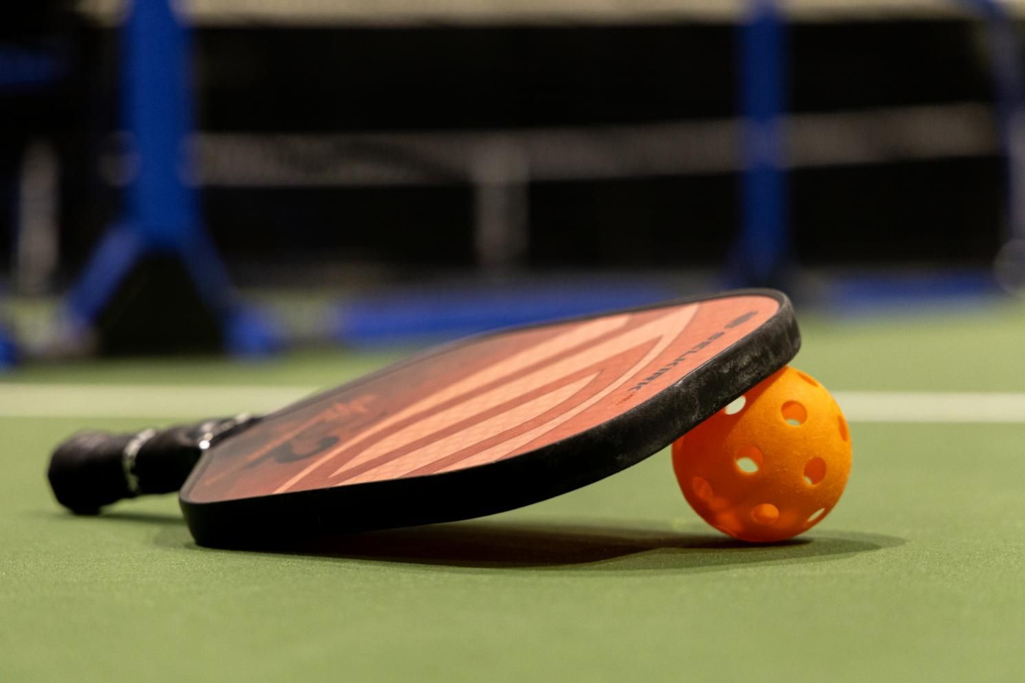 Pickleball paddle resting on an orange ball on a green court. A net is in the blurry background.