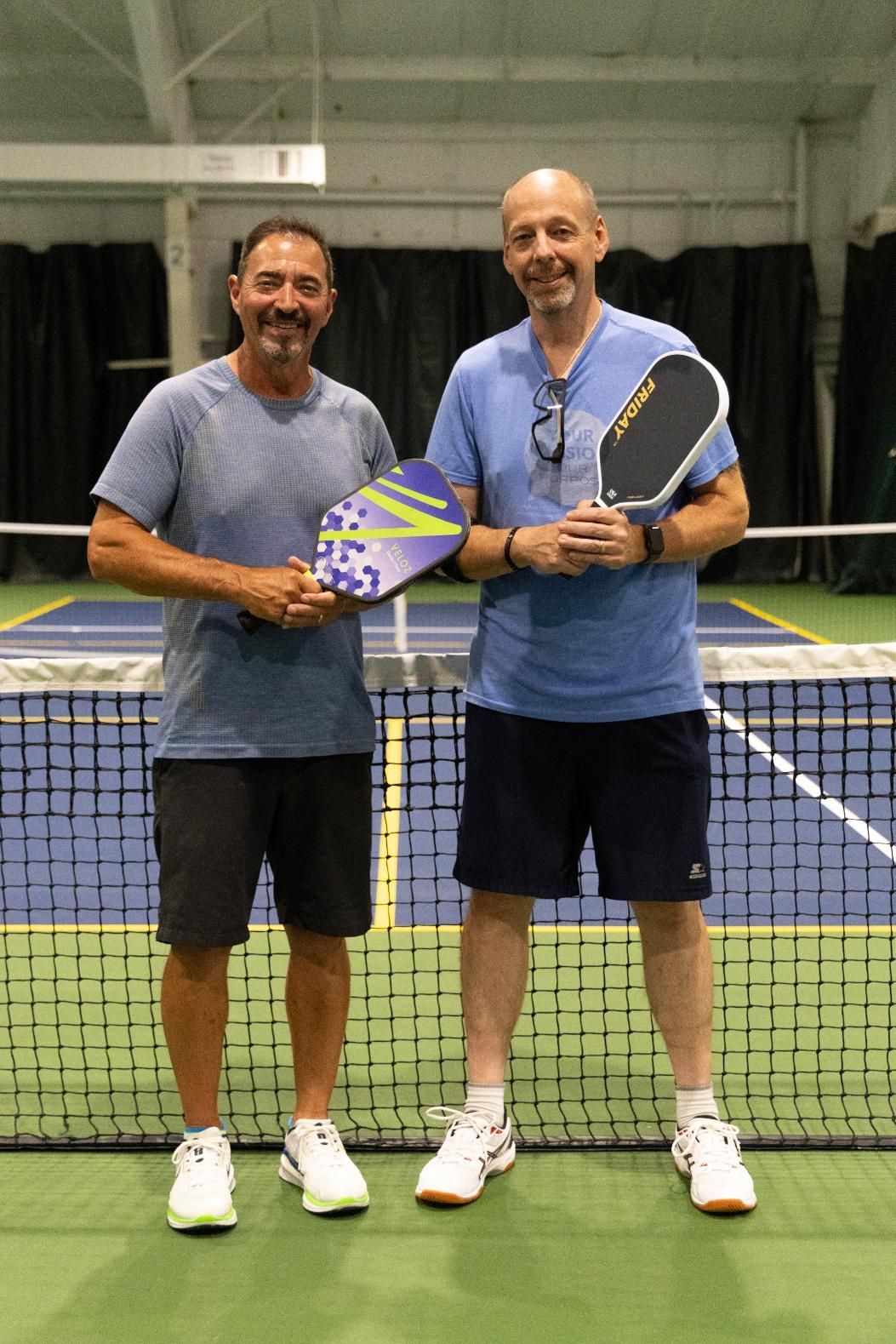 Two men stand on a pickleball court, holding paddles. They are smiling; the background is a net.