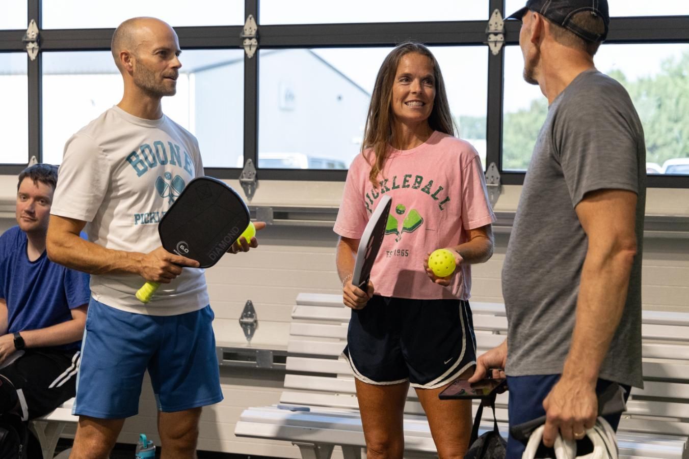 Three people with pickleball paddles and balls, talking inside a building.