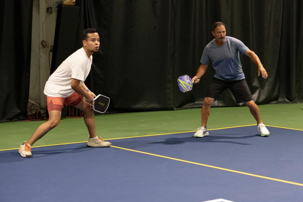 Two men playing pickleball on a blue court; one in white shirt and red shorts, the other in blue shirt and black shorts.
