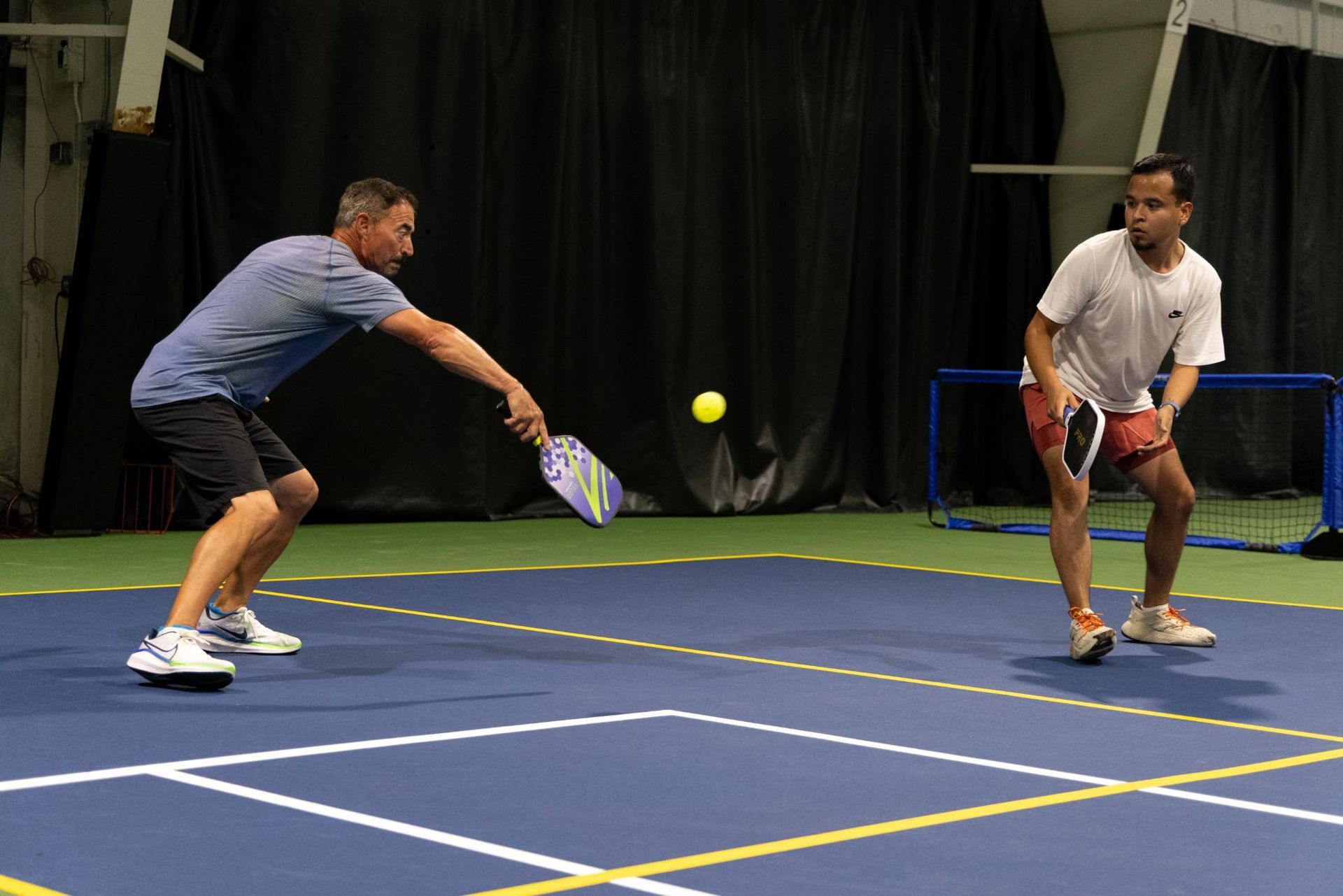 Man hits a yellow pickleball toward a boy on a blue court indoors. Both hold paddles.