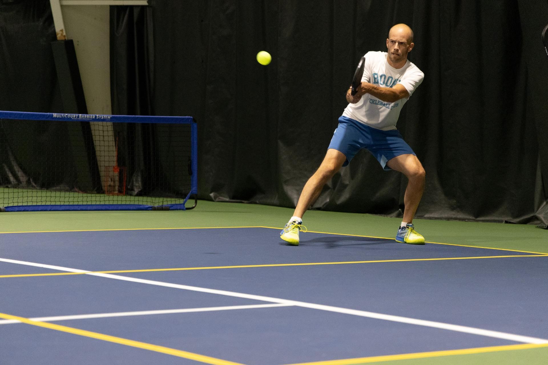 Man in white shirt and blue shorts playing pickleball indoors, hitting a yellow ball with a paddle.