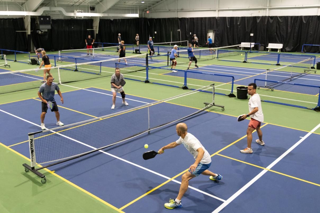 Pickleball game in indoor court. Players hitting ball over net, blue court with multiple playing areas.