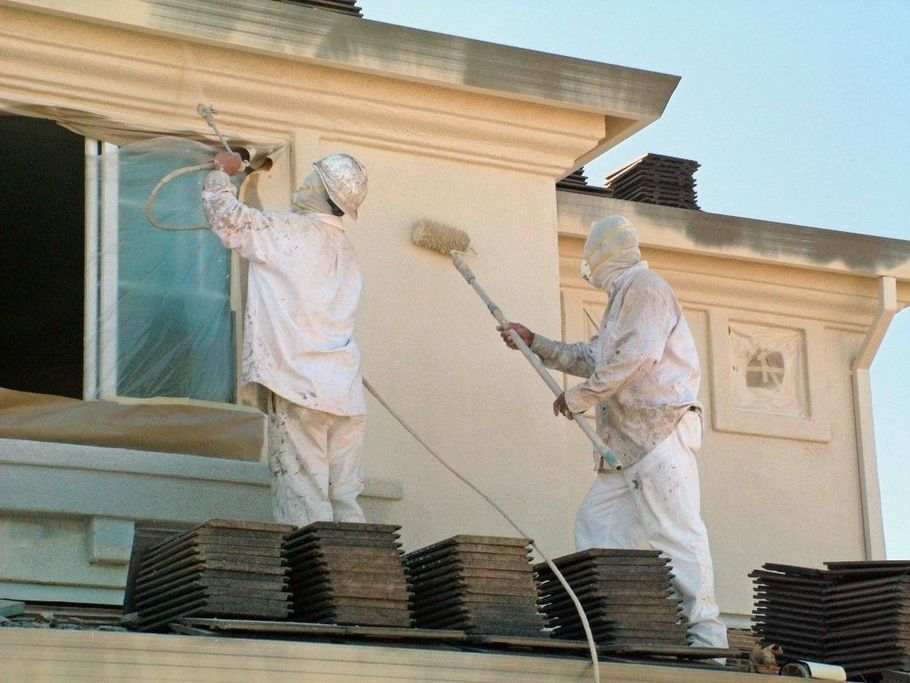 Two workers painting a house exterior, standing on stacked roof tiles with brushes and rollers.