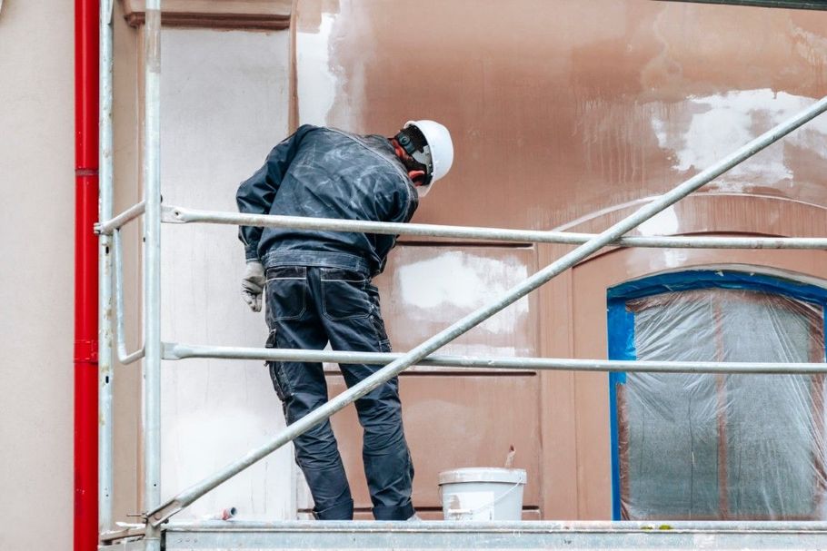 Worker painting a building facade from scaffolding, wearing a hard hat and safety gear.