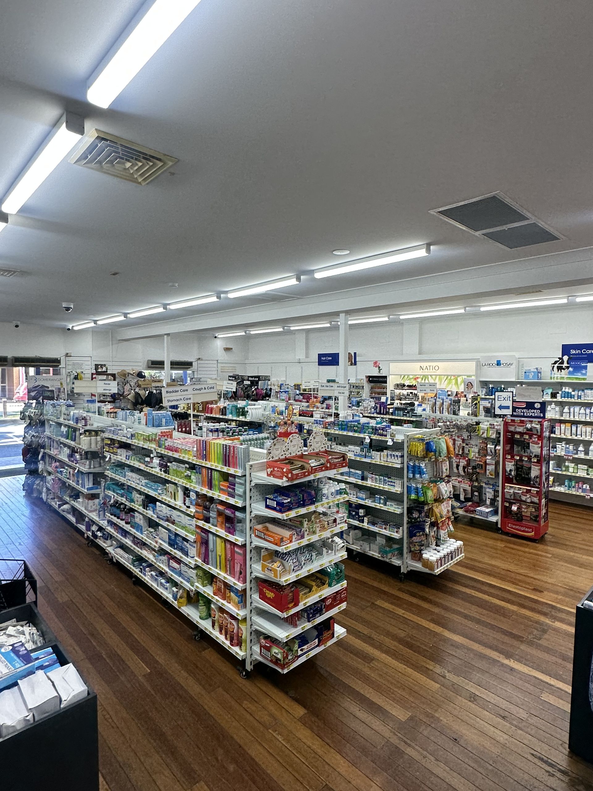 Interior of a pharmacy with shelves stocked with products, wooden floor, and fluorescent lighting.