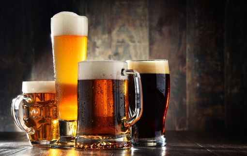 Four assorted glasses of beer on a wooden table, including a frothy tall lager and dark stout.