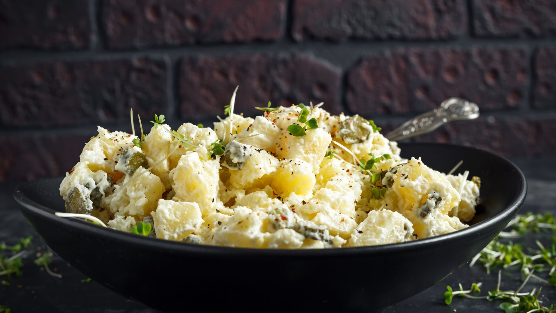Black bowl of potato salad with herbs and cheese on a dark table, with a spoon beside it