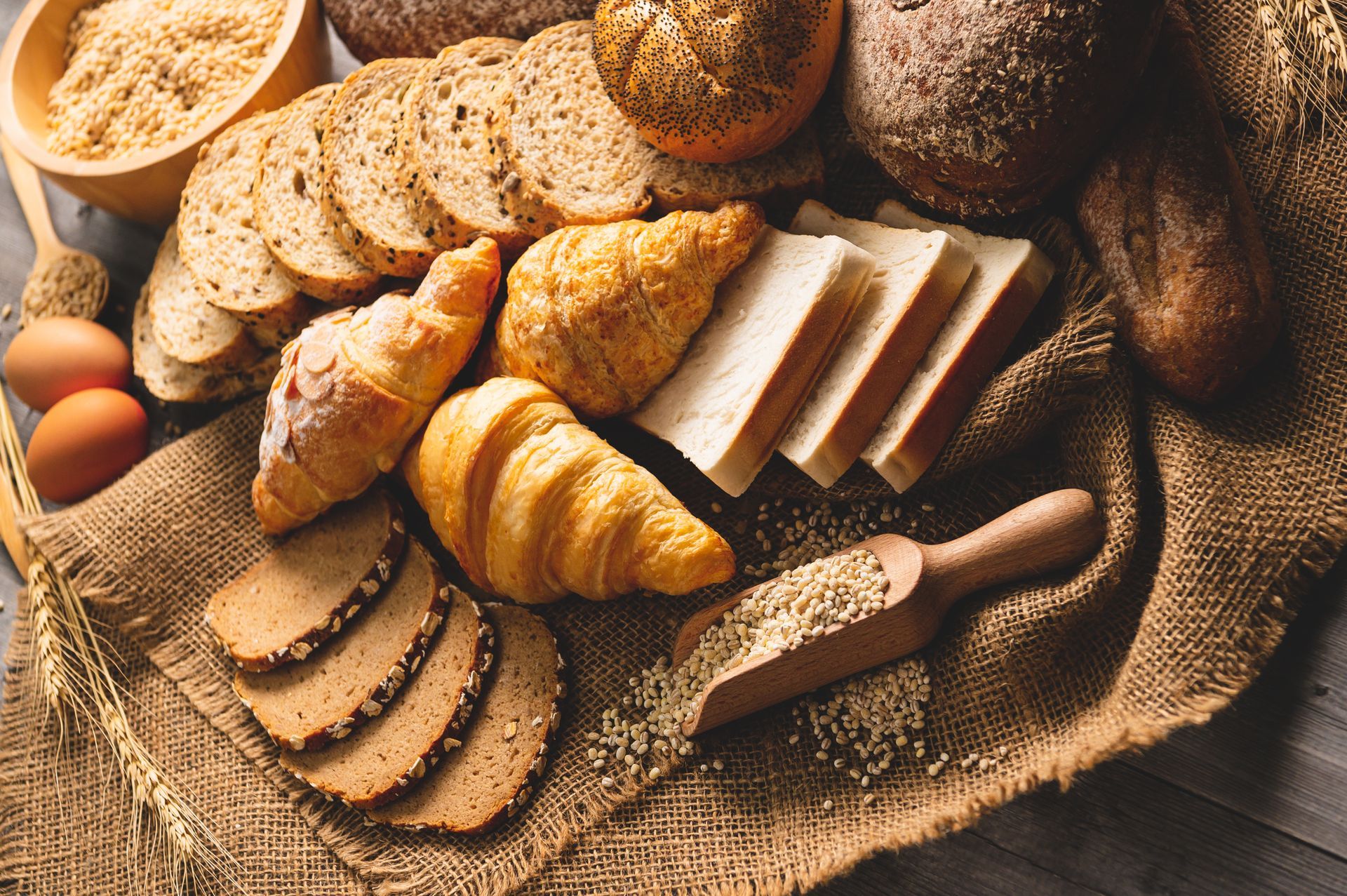 Assorted baked breads and croissants on burlap with a knife and eggs on a rustic table