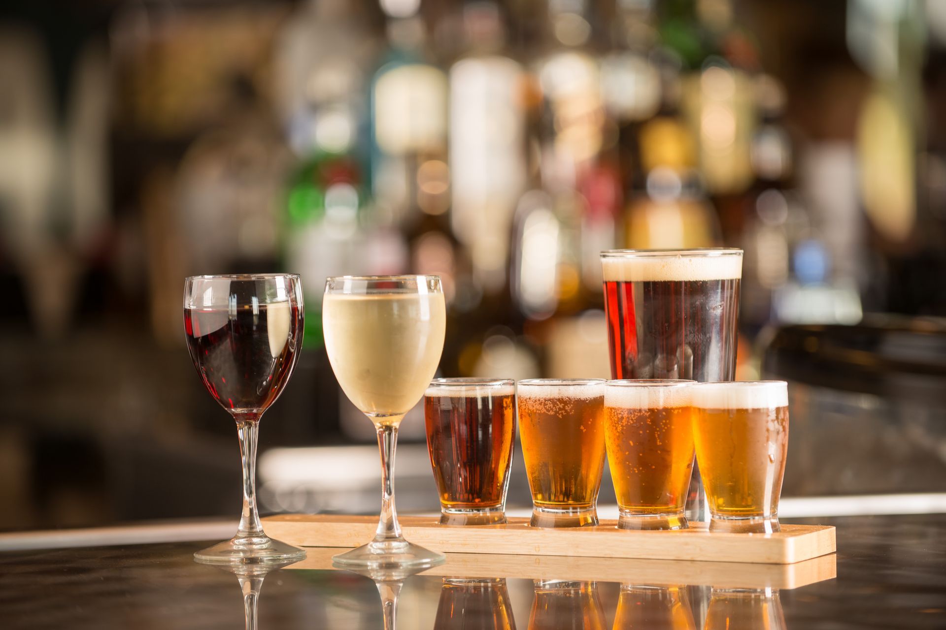 Assorted beers and wine glasses on a wooden tray at a bar with blurred bottles in the background