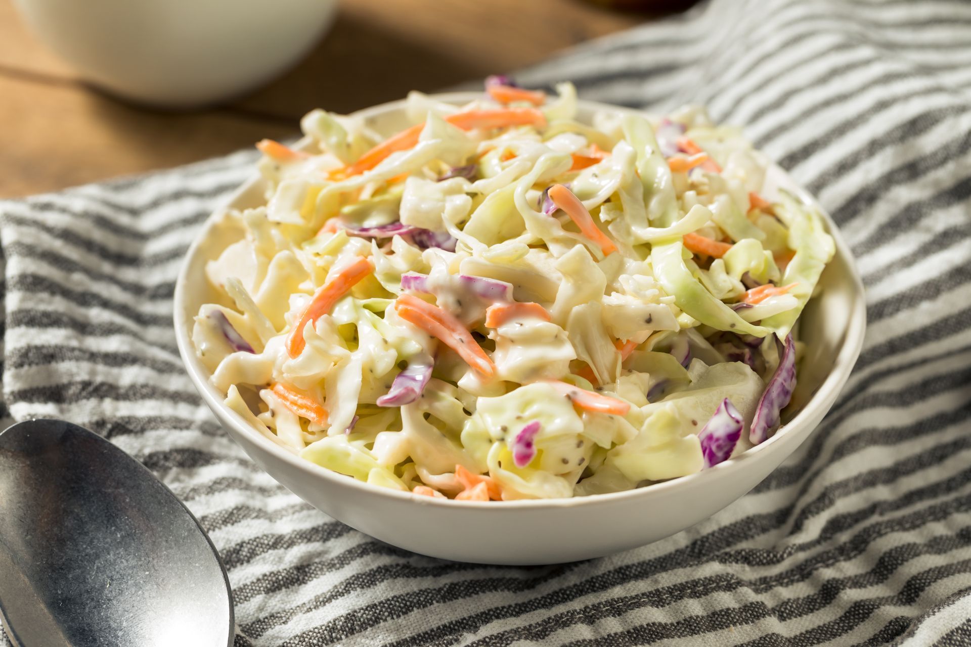 Bowl of creamy coleslaw with shredded cabbage and carrots on a striped cloth with a spoon
