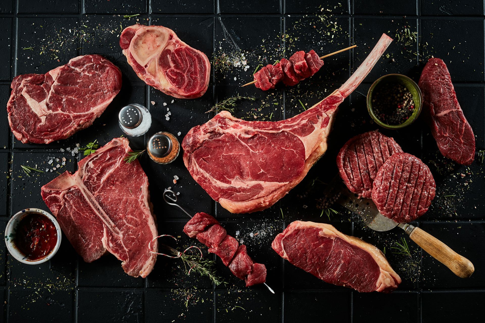 Raw assorted steaks and cuts of beef arranged on a dark slate surface with herbs and seasoning Raw assorted steaks and cuts of beef arranged on a dark slate surface with herbs and seasoning