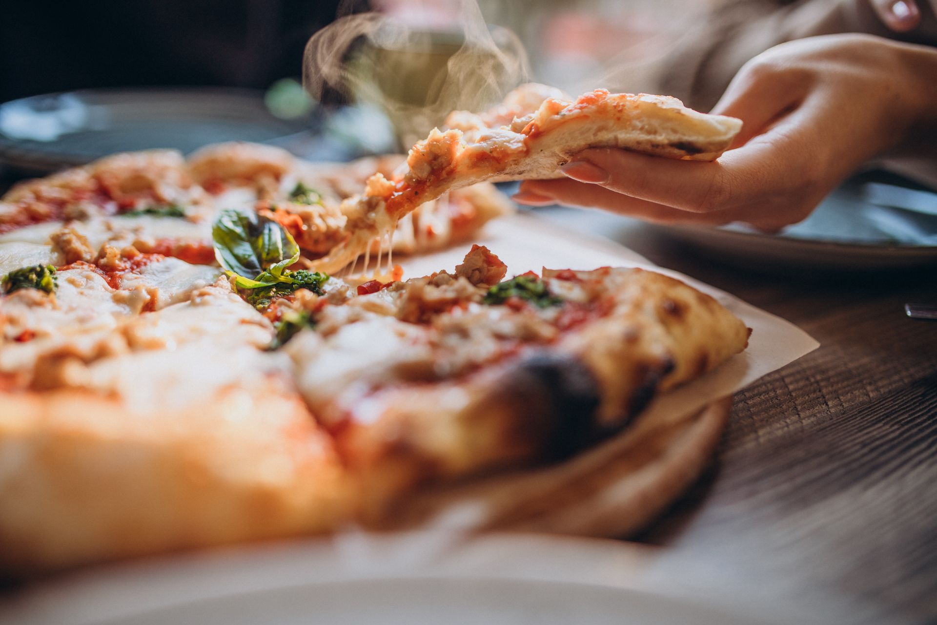 Hand taking a slice from a steaming cheese pizza on a wooden table