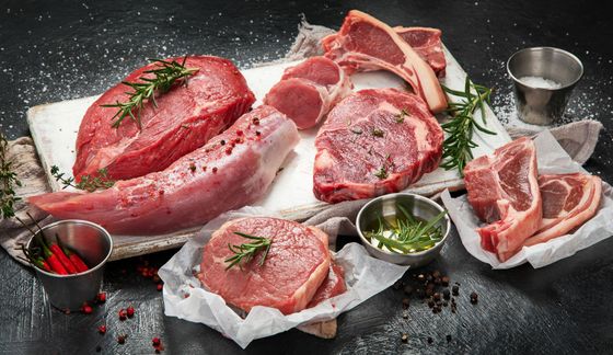 Assorted raw steaks and pork cuts on parchment with herbs and small bowls of seasoning on a dark surface