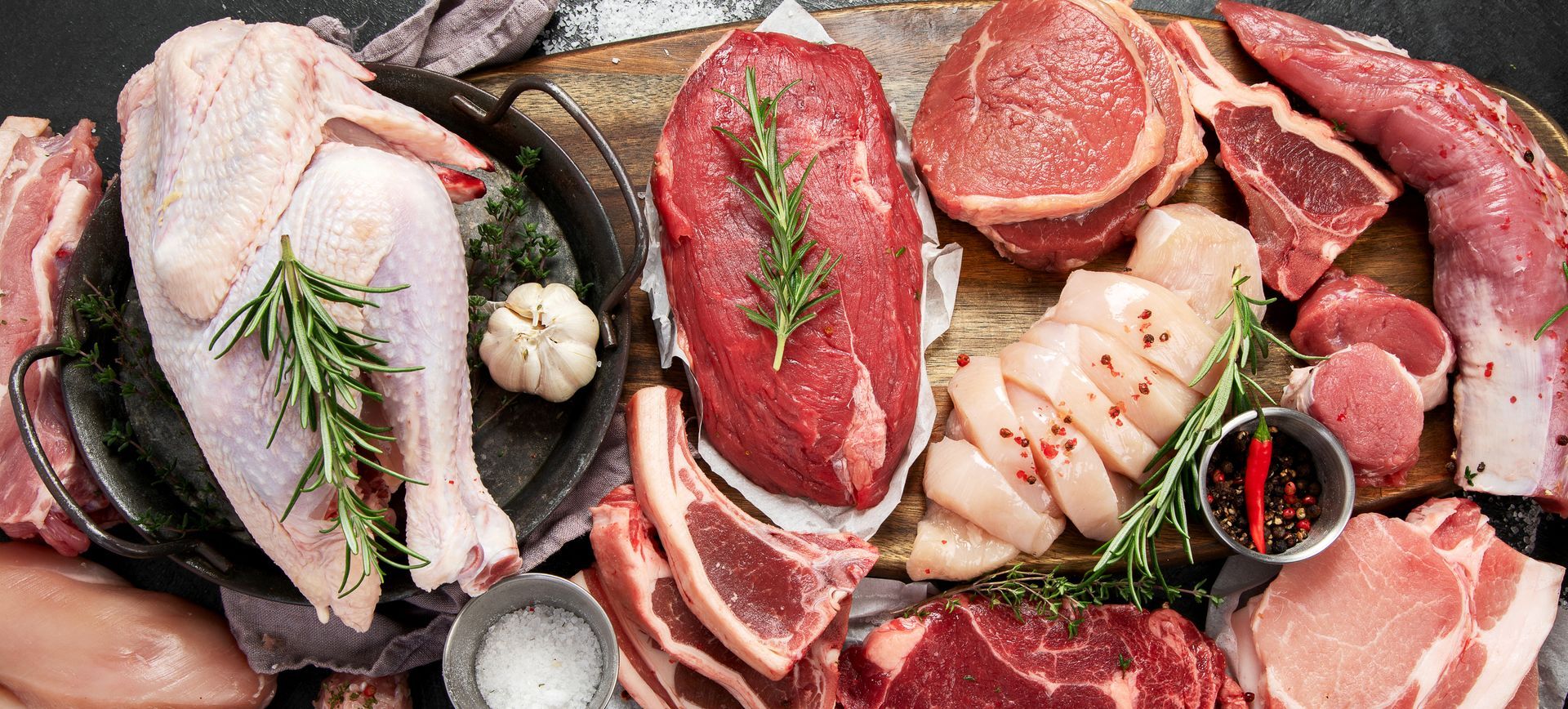 Assorted raw meats, steaks, chicken, and herbs arranged on a rustic wooden board.