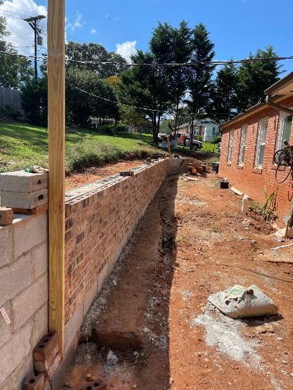 Brick retaining wall under construction beside a house, with dirt trench and wooden support posts.