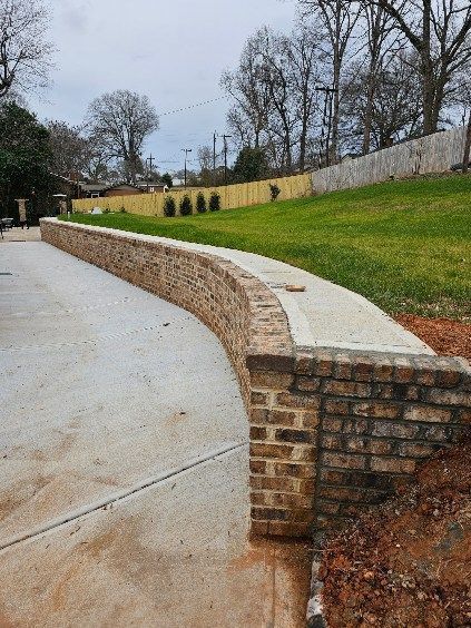 Curved brick retaining wall along a sloped grassy yard beside a concrete driveway under cloudy skies