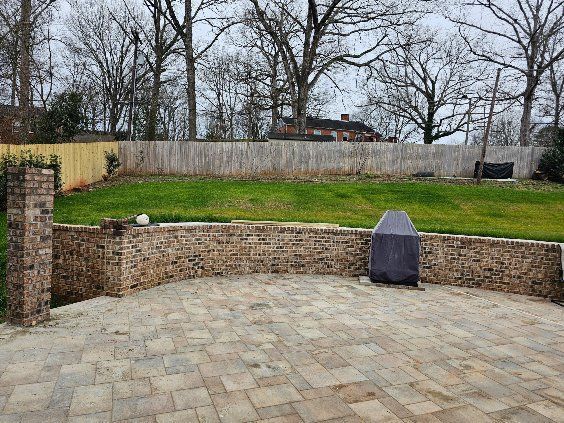 Paved patio with stone wall, grassy yard, bare trees, and a covered grill in the background