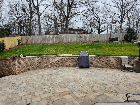 Backyard patio with stone seating wall, green lawn, leafless trees, and a covered grill center stage