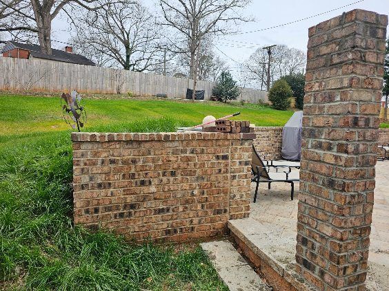Brick outdoor fireplace and low brick wall on a patio with grassy yard and trees behind.