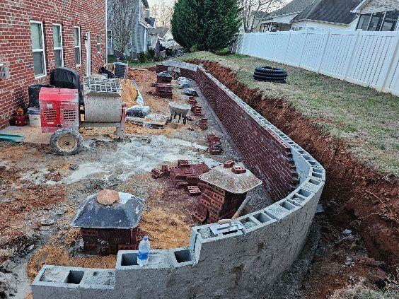 Curved concrete retaining wall under construction beside a brick house, with tools and materials in a backyard.