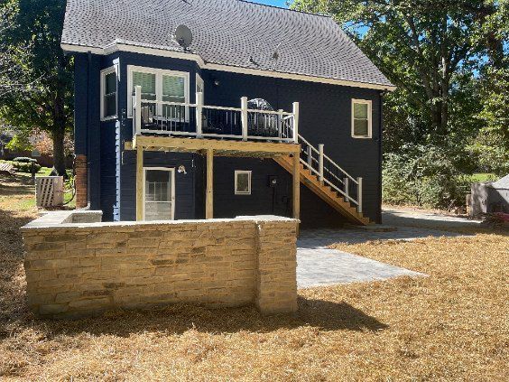 Backyard view of a dark house with a raised deck, exterior stairs, and a stone retaining wall.