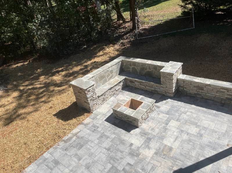 Stone patio with built-in fire pit and low retaining wall beside a wooded yard.