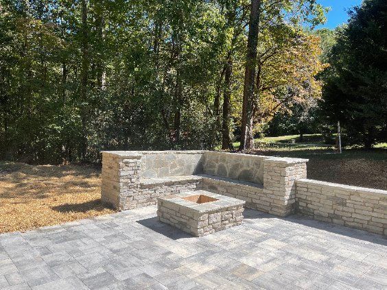 Stone patio with a built-in brick fire pit and benches, backed by trees and grass.