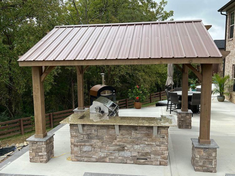 Covered outdoor grill pavilion with stone counter and brown metal roof in a backyard