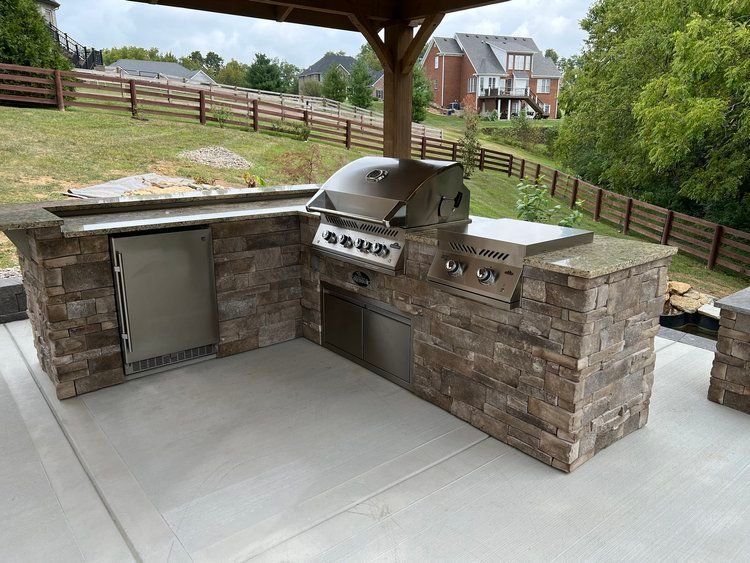 Outdoor stone barbecue grill and prep counter under a wooden pavilion in a backyard.