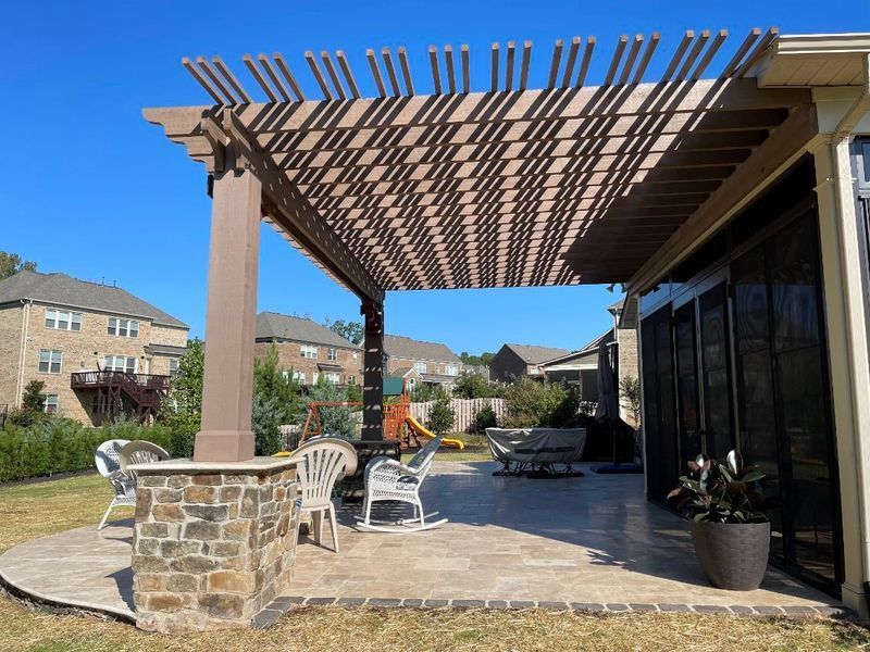 Patio with pergola, stone column, chairs, and potted plants beside a house under a clear blue sky