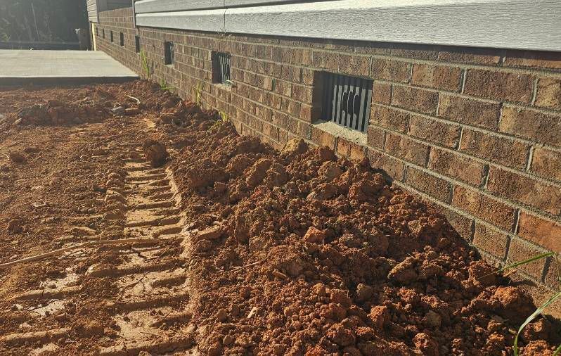 Freshly dug trench and piled red dirt along a brick house foundation line